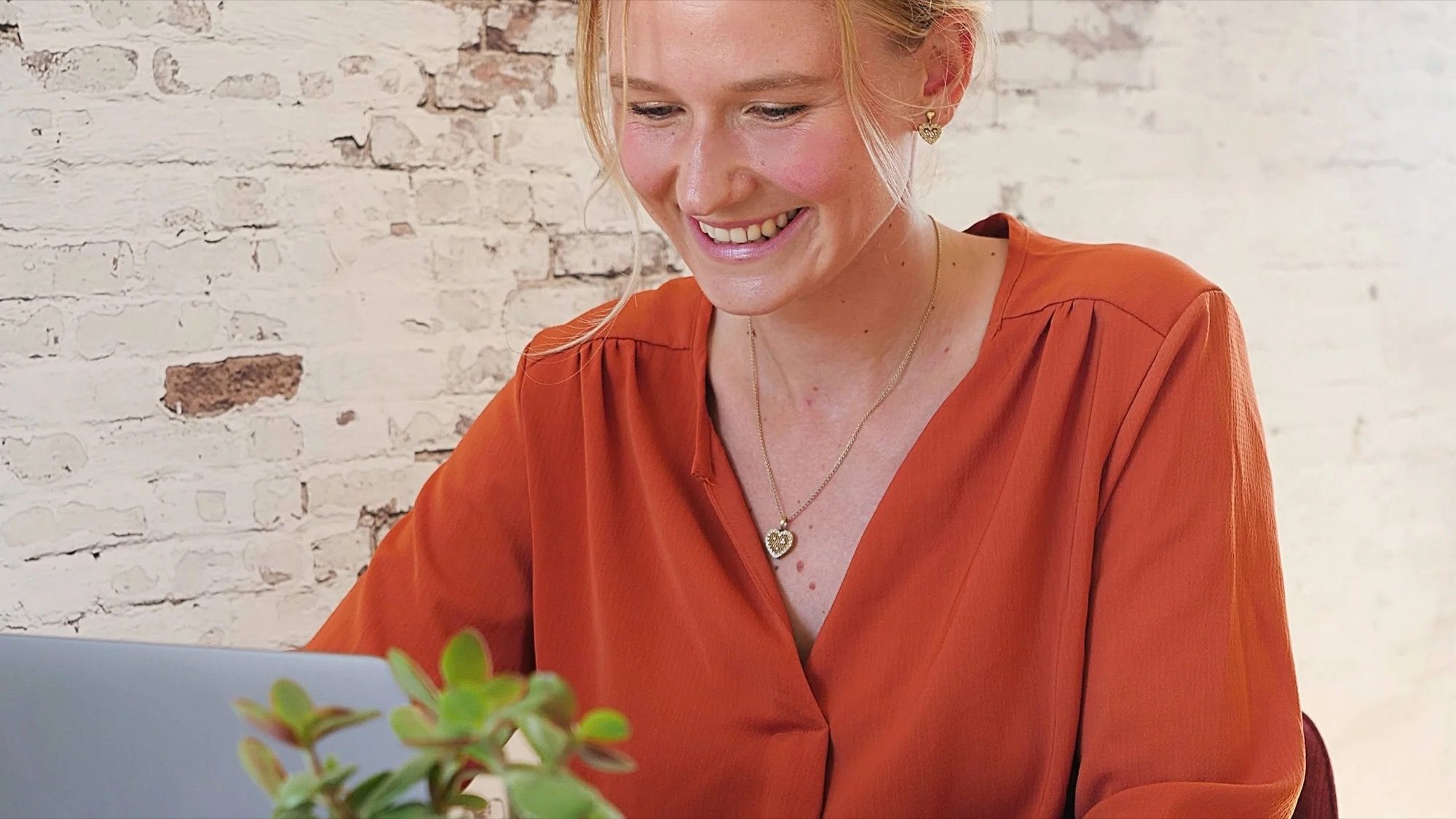 Alizé Foveau, en chemisier orange, sourit devant son ordinateur portable, adossée à un mur de briques peintes.