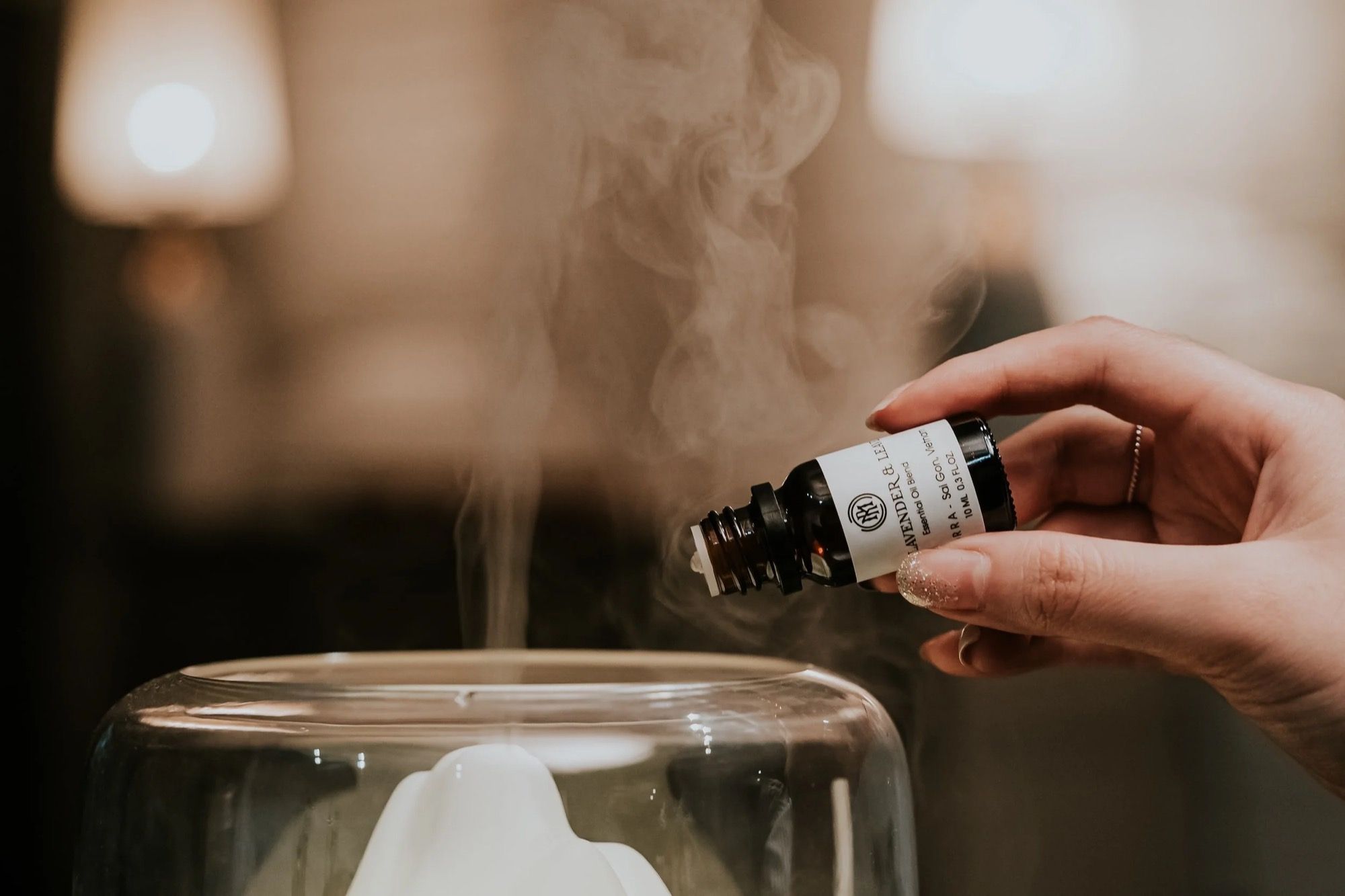 Une main approche un flacon d’huile essentielle d’un diffuseur d’où s’élève un filet de vapeur.