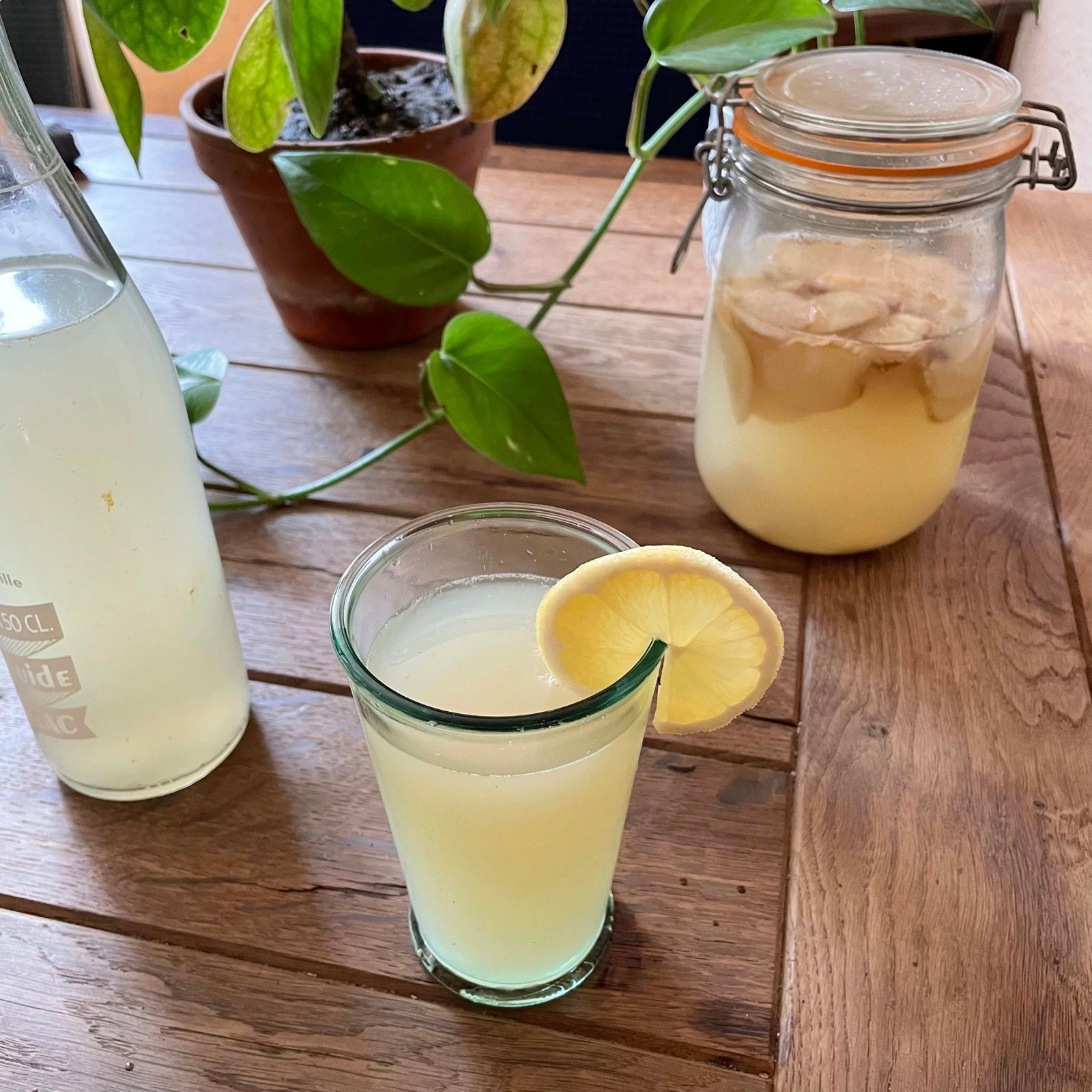 Un verre de ginger beer maison avec une rondelle de citron, la bouteille et le bocal de gingembre à côté.